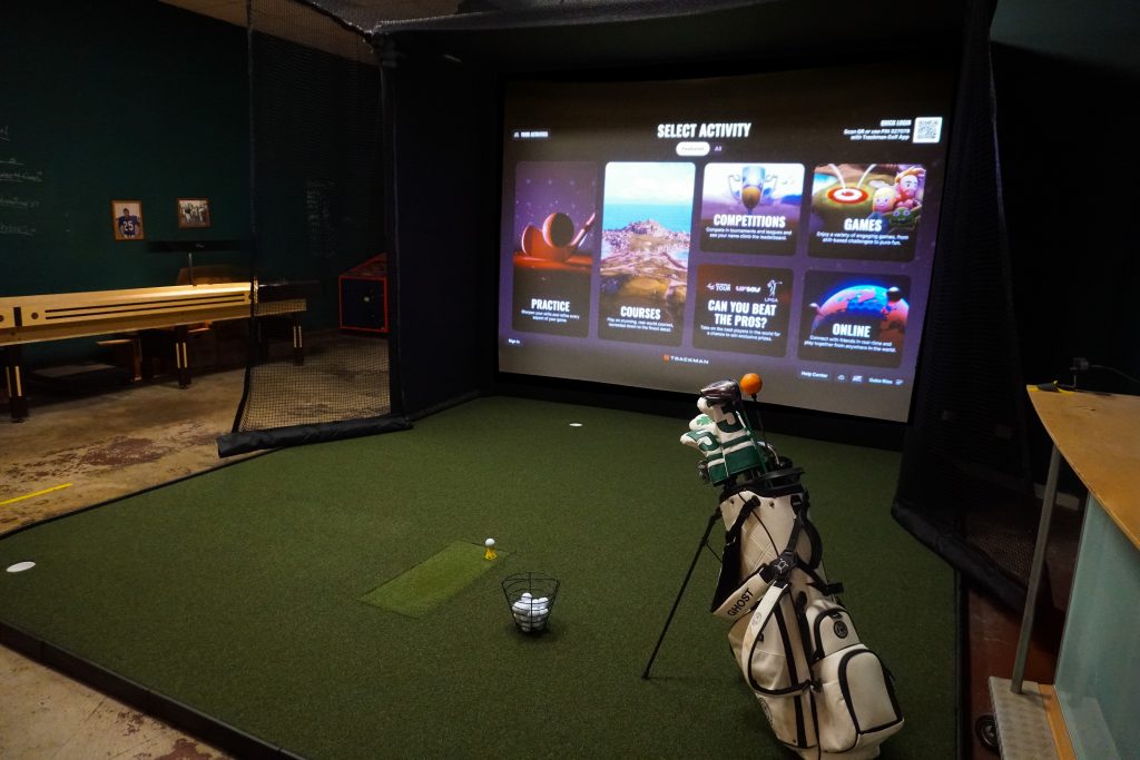 Golfer practicing in an indoor golf simulator with exposed brick walls and a projected fairway.